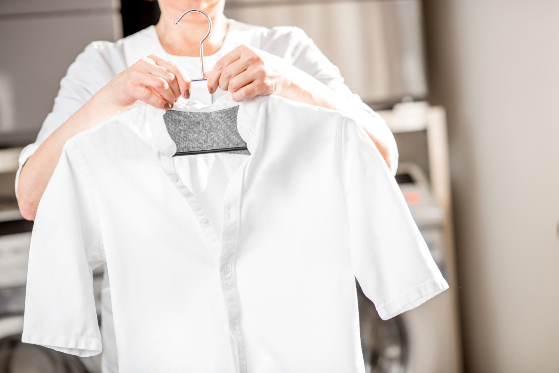 A female employee in a dry cleaning shop, wearing a uniform with a yellow scarf, holds up a white shirt on a hanger, smiling at the camera.